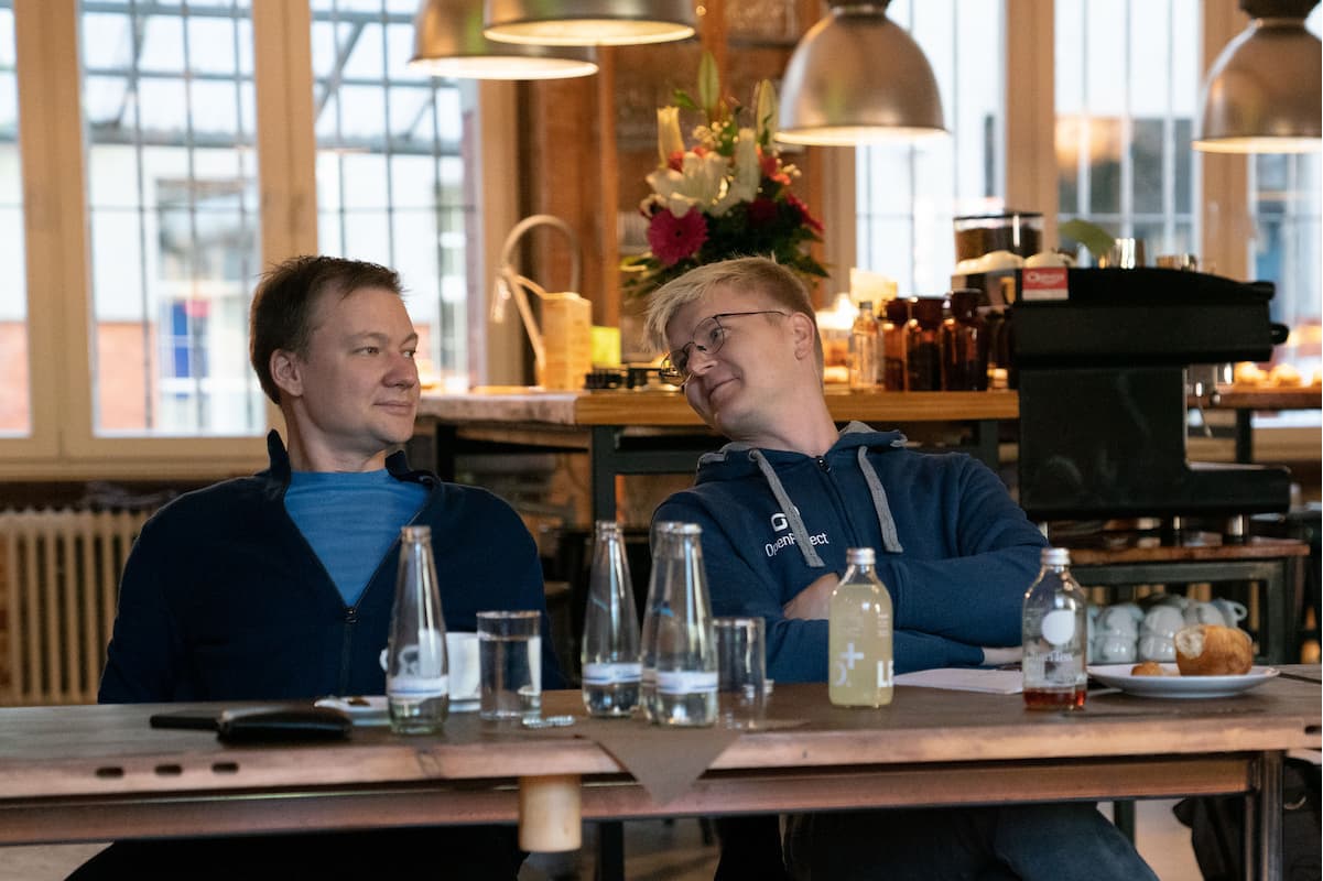 Dos hombres sentados en una mesa de comedor de madera con comida y bebida, enfrascados en una conversación.